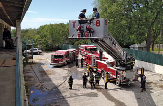 A newly promoted apparatus driver learns from the senior truck firefighter that, depending on how the apparatus is spotted and how the aerial device is deployed, the 105-foot platform will still not reach some places, the scrub areas.