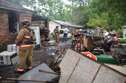 (2) A view along the D side shows several vehicles and a utility trailer blocking the driveway. A guard dog chained to the rear bumper of the trailer forced firefighters to advance a hoseline and move equipment through the row of bushes to the right. (3) A closer view of the D/C corner where personnel made the transitional attack into the fire room. Waist-high piles of debris in the carport area stopped the crew in an attempt to make the initial push into the dwelling. (4) At the C/D corner, an overwhelming amount of debris blocked the window and the door. These conditions could have prevented the occupants from escaping and could have hampered any rescue effort or emergency egress of firefighters.