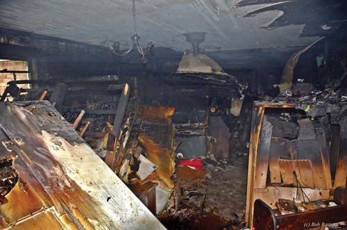 (5) A view through the A side door shows the piles of debris throughout the room. Communicating these conditions immediately to Command resulted in a change of strategy to an aggressive defensive attack from the D/C corner. Next, a ventilation fan was placed in service at the A side entrance prior to personnel moving back into the structure for extinguishment. The refrigerator to the left was partially behind the door and had to be knocked over to gain entry. 