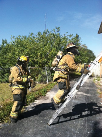 (2) The search position firefighter (FF) climbs the ladder as the window position FF dons face piece and gloves. Note the search position FF is on air and is carrying the halligan up to use during his search. Prior to climbing the ladder, the search position FF had hung the six-foot roof hook on an upper rung so he can use it to vent the window. 