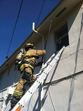 (3) The search position FF ventilates the window from a ground ladder. Note the modified leg lock using a simple knee press to a higher rung. This member must be on air and ready to make immediate entry. The member is positioned under the windowsill for safety should the fire react violently on ventilation. The tips are positioned at the sill, and the ladder is at an exaggerated climbing angle. 