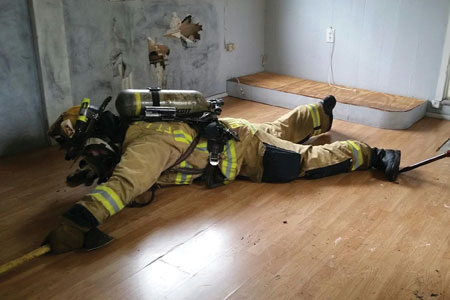 (8) The search position FF then lies flat on the floor under the smoke to get a layout of the room and to locate the bedroom door. The roof hook to the right of the firefighter’s foot provides a reference point toward the window’s location.