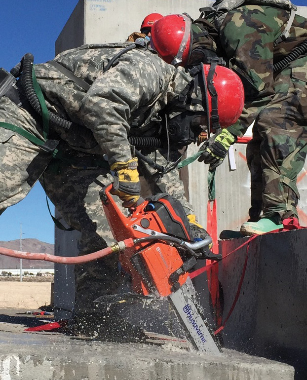 During a Nevada training exercise, rescuers use a concrete chain saw with a diamond blade to cut through the full thickness of the concrete and rebar. The rescuer operating the saw has a webbing harness to prevent himself from falling into the hole he is cutting.