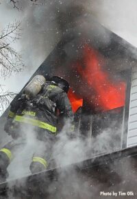 Flames shoot out from an upper story as a firefighter operates on the roof