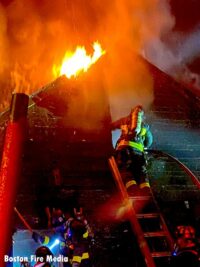 Flames exposed from roofline as Boston firefighter on a ladder works at house fire