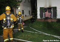 LAFD firefighters advance a ladder between structures while U.S. flag hangs in window