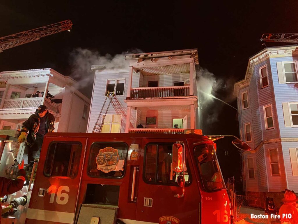 Firefighter on a fire truck with fire building behind and two aerials in use in Boston