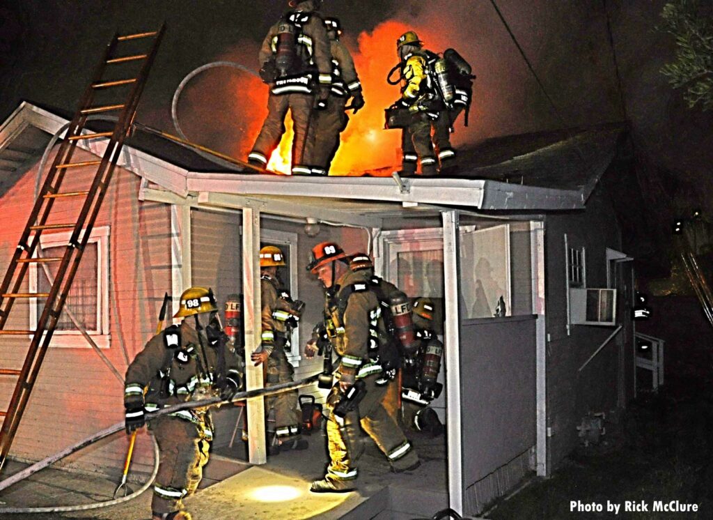 Firefighters on a roof as other members work a hoseline and advance it into the home