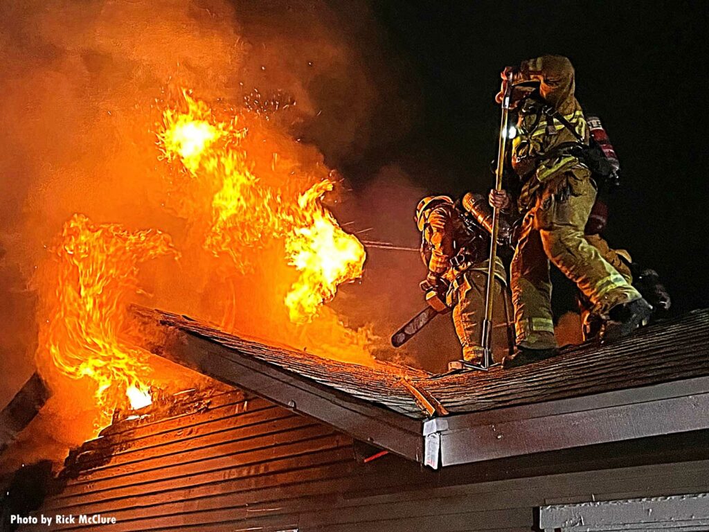 One firefighter a saw and another with a hook as fire vents through the roof