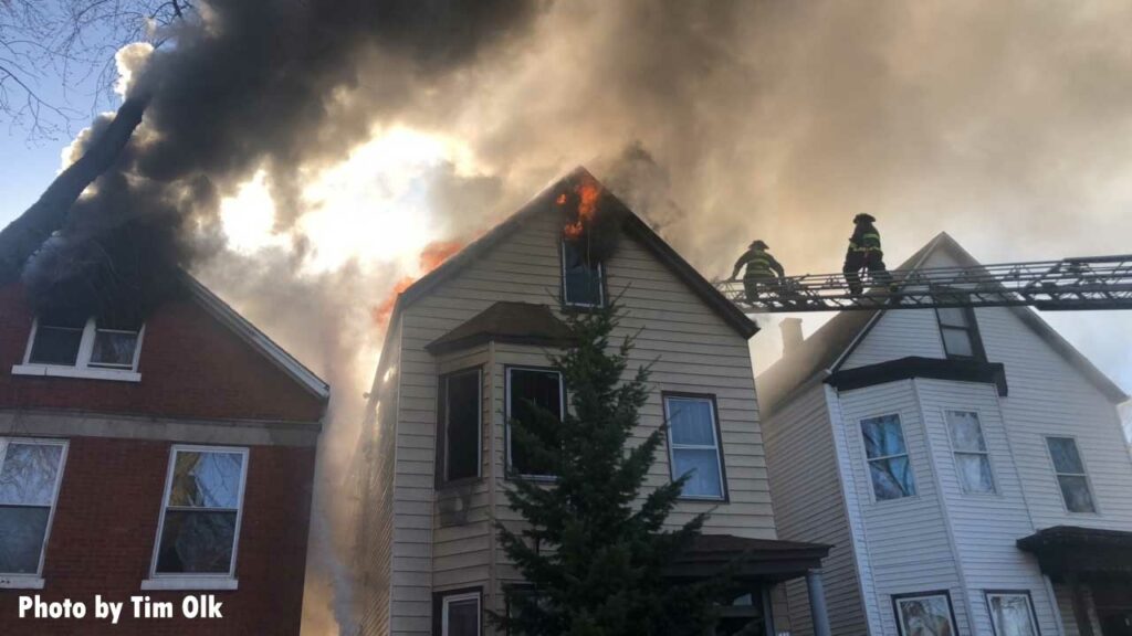 Smoke showing from one home with smoke and flame showing from another and Chicago firefighters on an aerial ladder