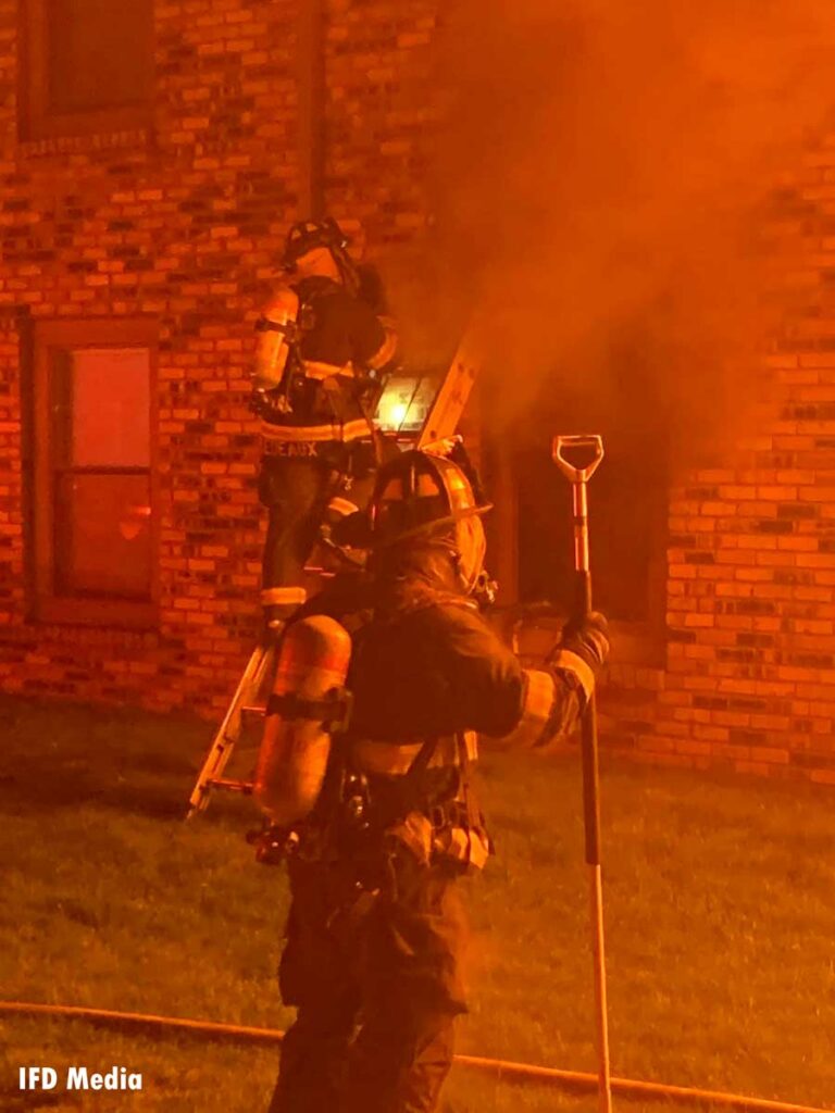 Firefighter ascends a ladder at the scene of a fire in Indianapolis while another firefighter is in the foreground with a hook