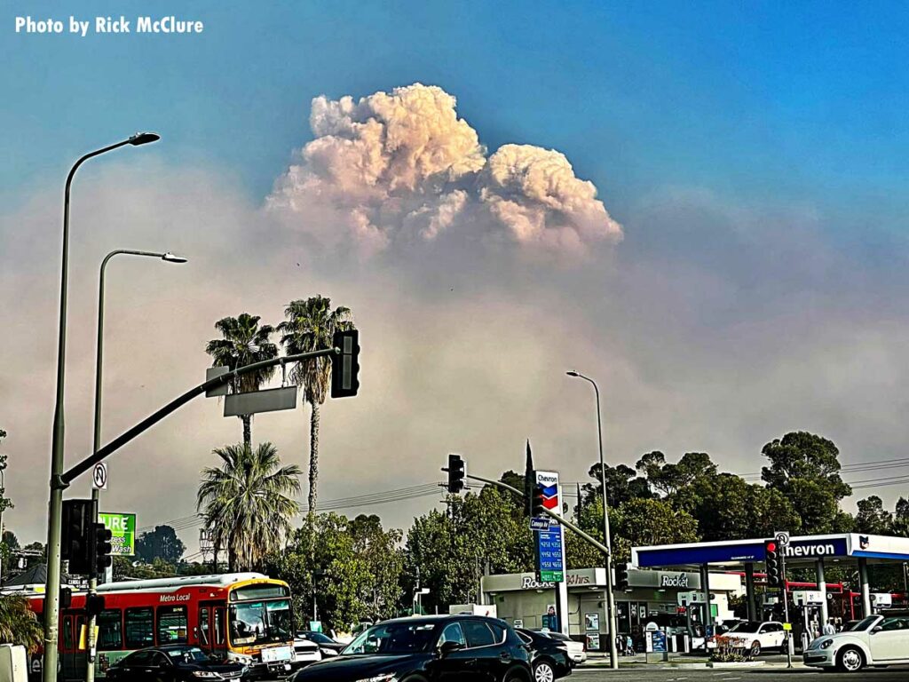 A street with a gas station and palm trees in the foreground with a plume of smoke from the Palisades Fire rising in the distance