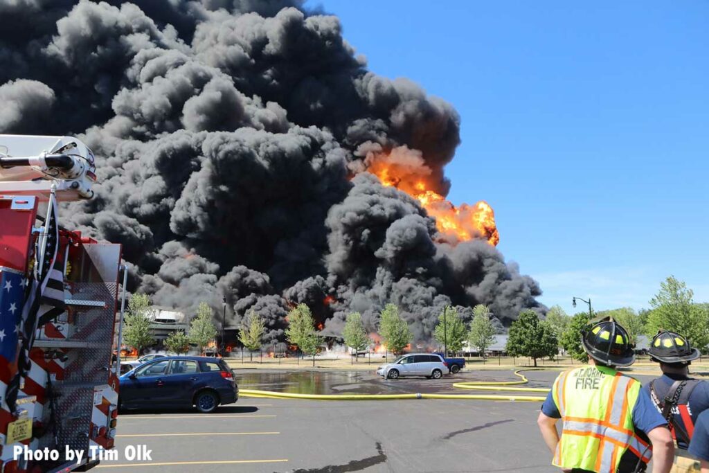 Huge flames and smoke shoot into the sky with firefighters in the foreground