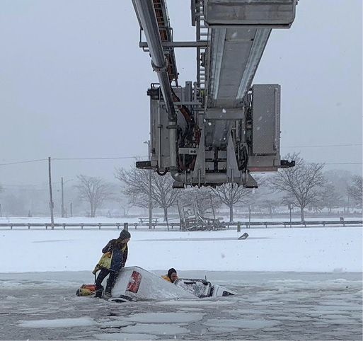 The tower ladder’s basket is maneuvered out and over the rear cargo area of the sinking truck as rescue swimmers attempt to gain entry to rescue the remaining male occupant in the passenger seat. The female driver stands at the rear of the truck’s bed.