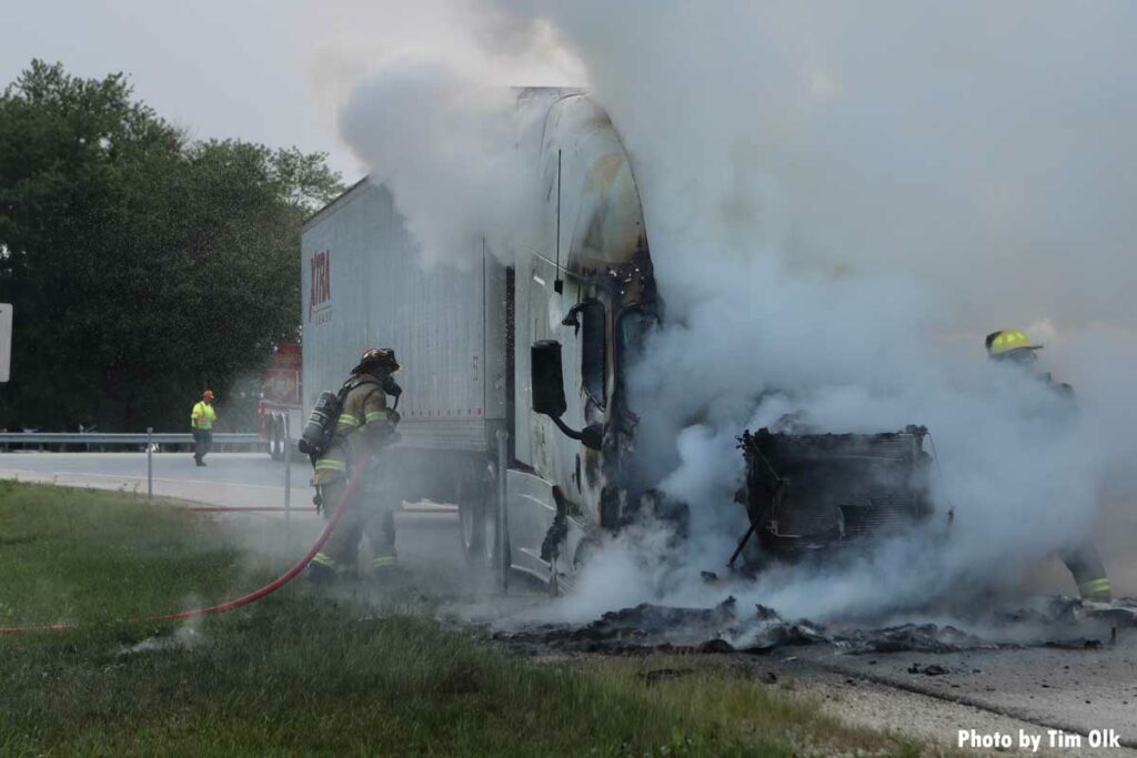 Smoke wafts from a burning semi cab as firefighters advance a hoseline