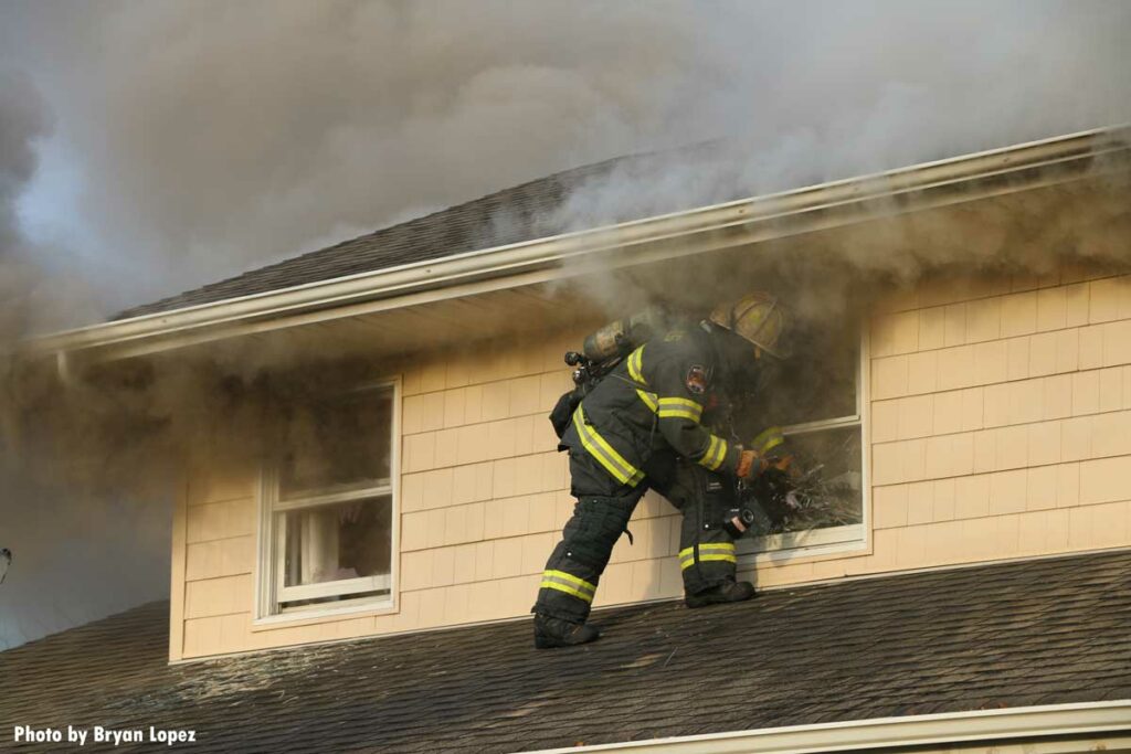 Firefighter clears window on second floor with smoke wafting from underneath