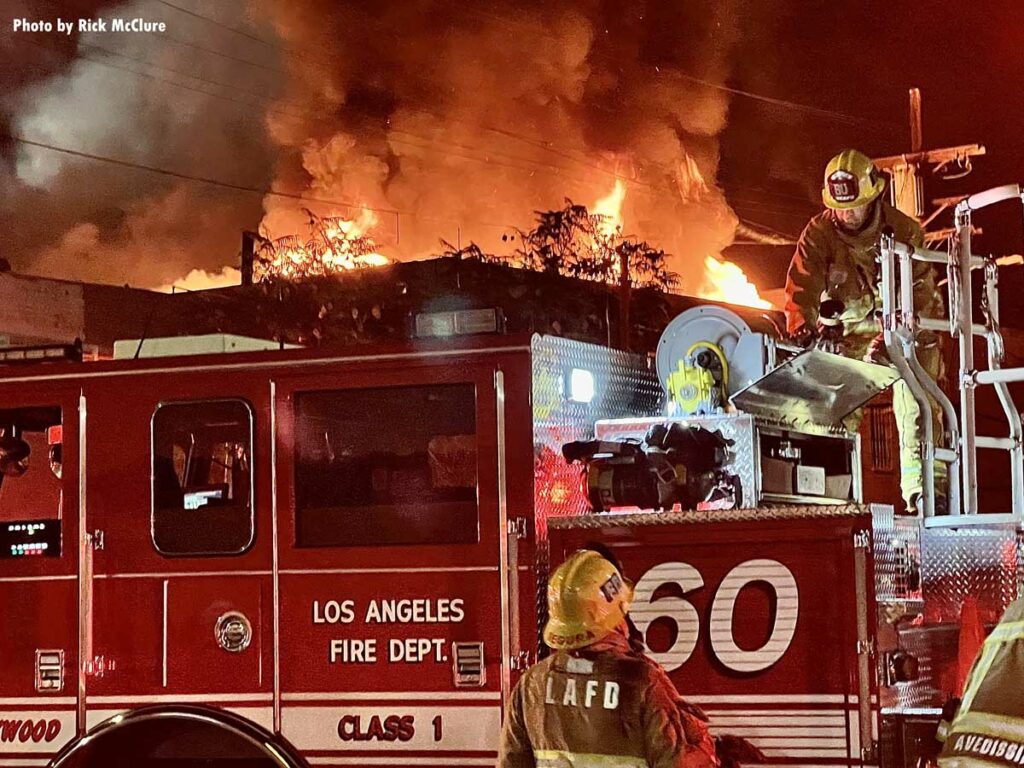 LAFD firefighters on top of engine with burning commercial structure in background